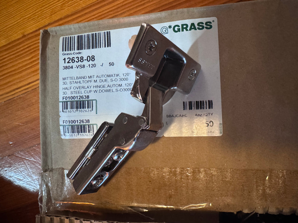 A metal cabinet hinge mounted on its original brown cardboard packaging with a white label. The brand name 'Grass' is clearly visible on the label.